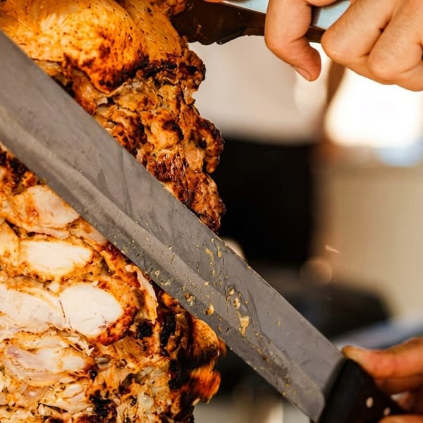 A chef's hands expertly carving juicy, seasoned chicken from a vertical rotisserie skewer.