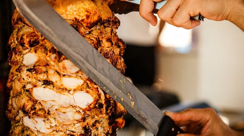 A chef's hands expertly carving juicy, seasoned chicken from a vertical rotisserie skewer.