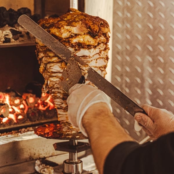 A chef in gloves carves meat from a large vertical rotisserie with glowing coals below.