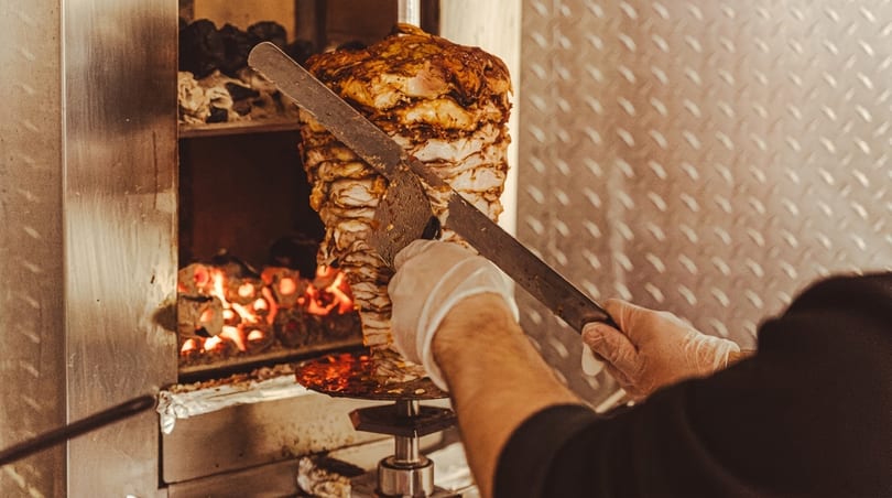 A chef in gloves carves meat from a large vertical rotisserie with glowing coals below.