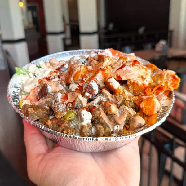 Hand holding a foil bowl of rice, gyro meat, spicy chicken, and two sauces in a restaurant.