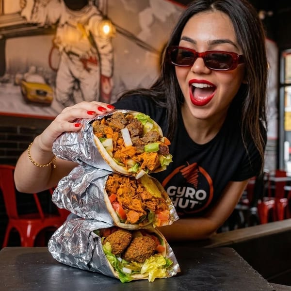Excited woman in sunglasses holding three stacked gyros from Gyro Hut.