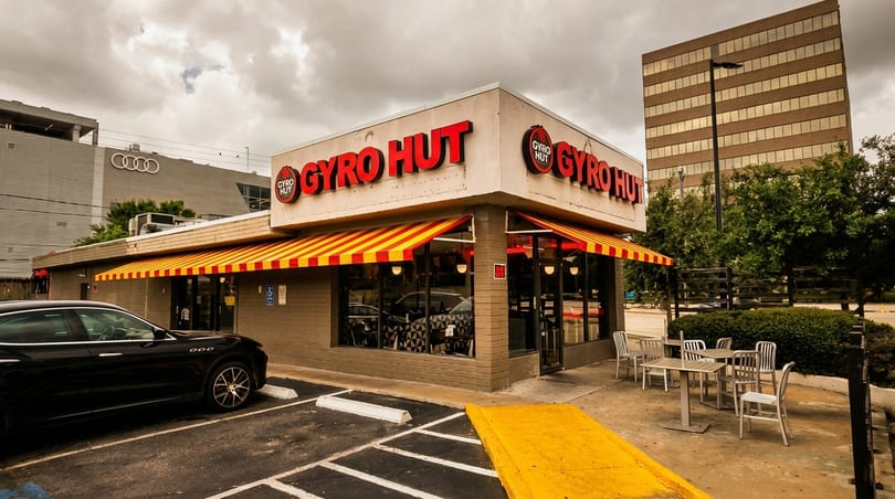 Exterior of Gyro Hut restaurant with red and yellow striped awnings, outdoor seating, and a parked car.