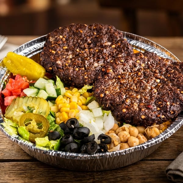 Two seasoned burger patties in a foil bowl with corn, chickpeas, olives, and chopped vegetables.