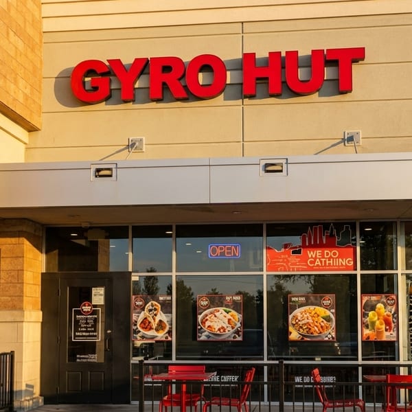 Exterior of Gyro Hut restaurant with red sign, glass windows displaying food, and red outdoor seating.