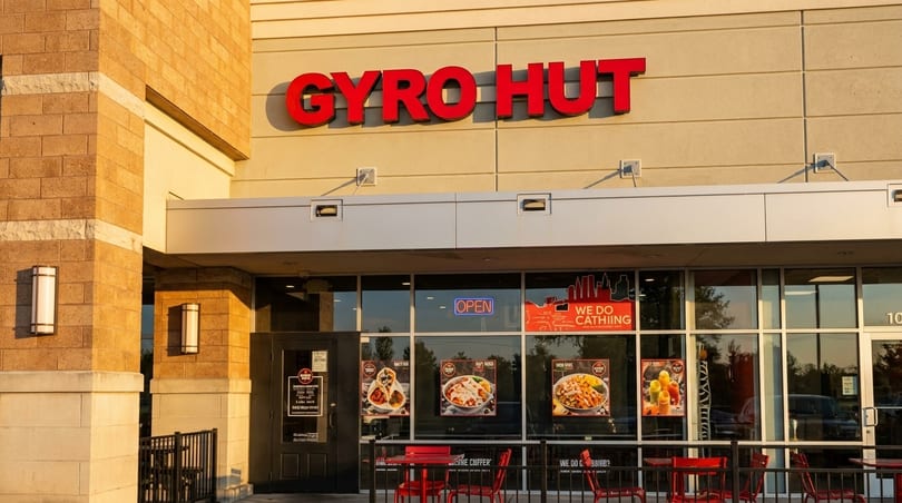Exterior of Gyro Hut restaurant with red sign, glass windows displaying food, and red outdoor seating.