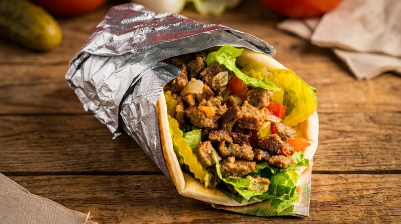 A beef shawarma wrap with lettuce, tomatoes, and pickles, partially unwrapped from foil on a wooden table.